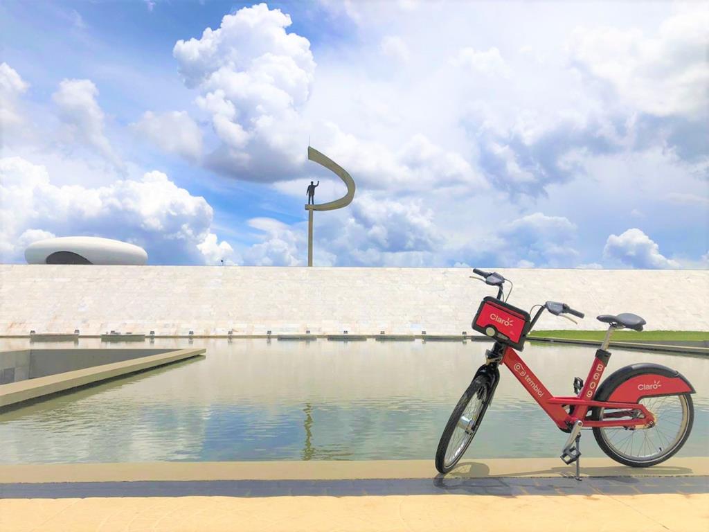 Bicicleta da tembici, estacionada em um local deserto, próximo a um lago e um monumento ao fundo.