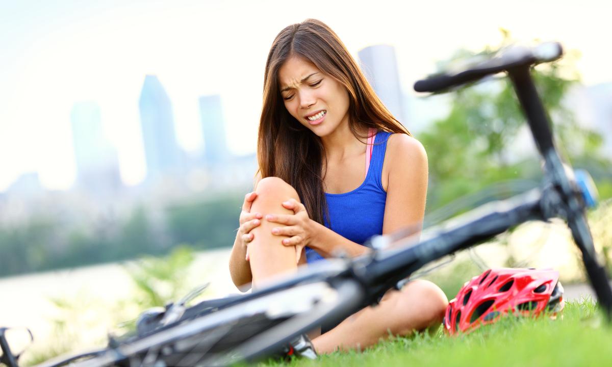 mulher com as mãos em seu joelho, com expressão de dor, e sua bicicleta ao chão em sua frente
