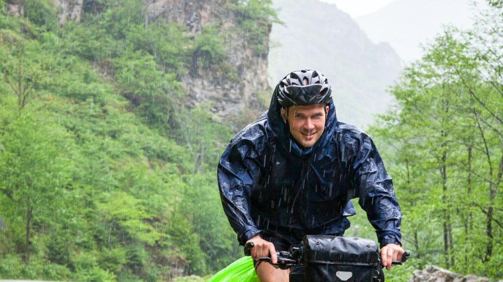 homem pedalando na chuva com capa, capacete de proteção e demais acessório de segurança