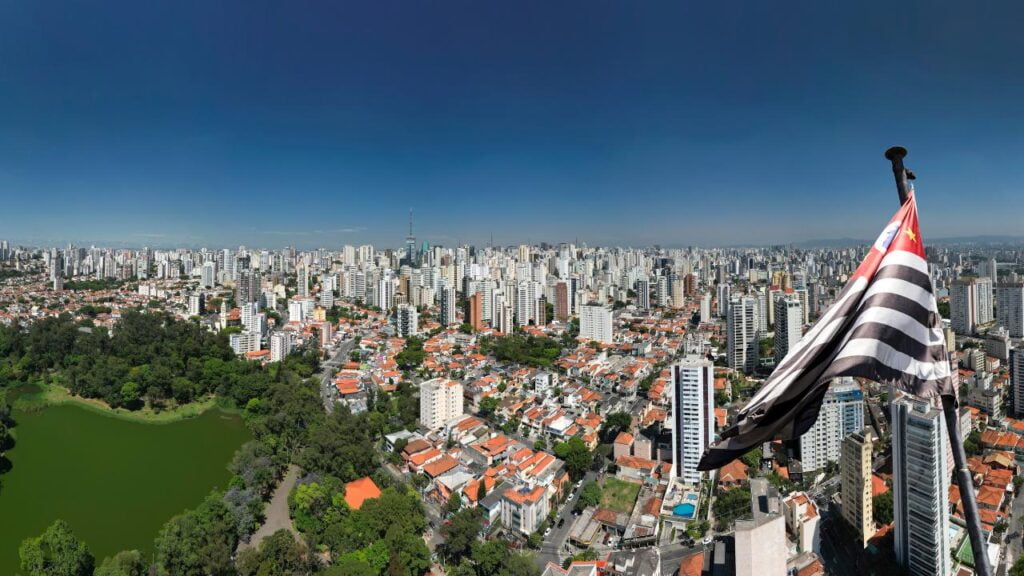 imagem aérea da cidade de São Paulo, uma bandeira do ESTADO a direita e um parque a esqueda da imagem. Parques são excelente opção de diversão de graça em SP.