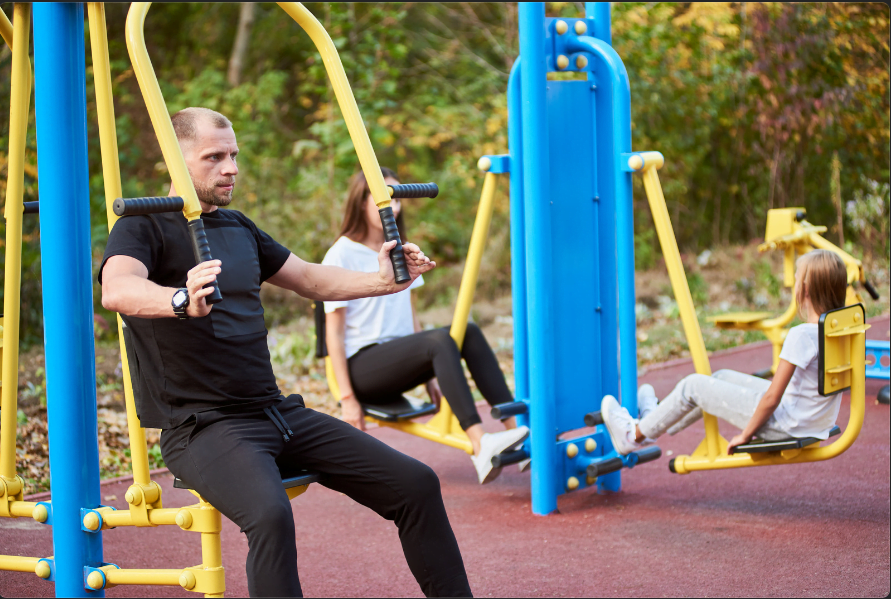 Homem praticando atividade física em parque com aparelhos utilizados para alongamento dos músculos e, mais especificamente, dos ombros e braços.